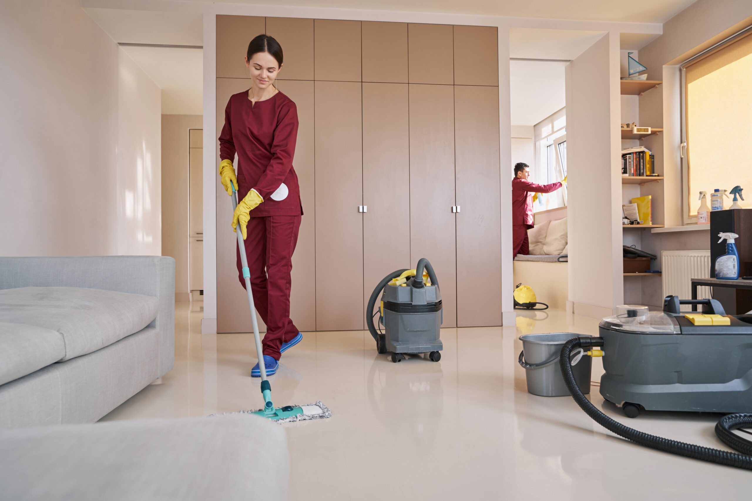 Smiling female janitor washing floor with microfiber dust mop while her colleague cleaning window
